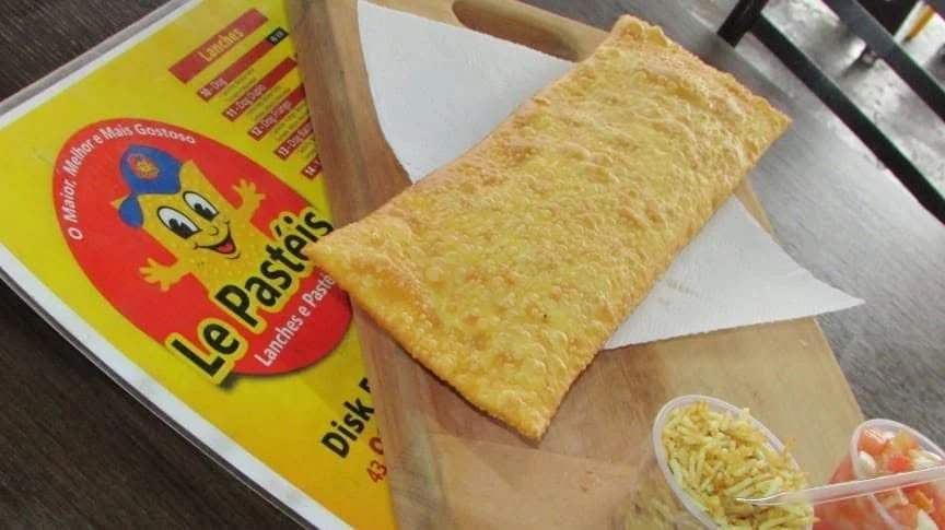 Pastelão dourado na tábua, com papel, potinhos de batata palha e vinagrete e o cardápio impresso ao lado
